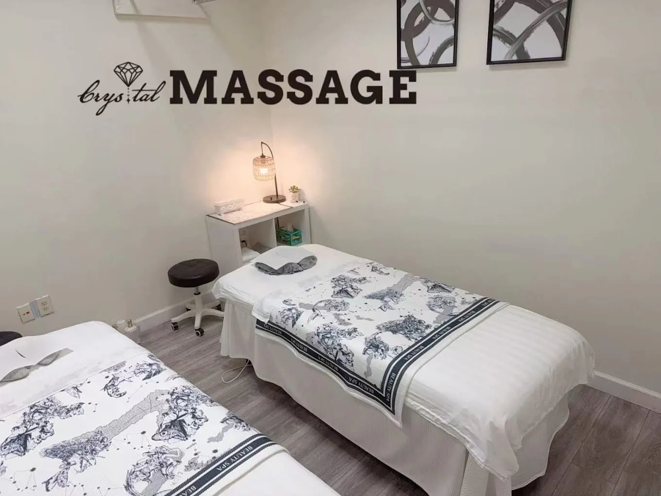 Treatment room with two massage tables made up in white linen, a lamp on a console and the Crystal Massage name on the wall, Wichita, KS